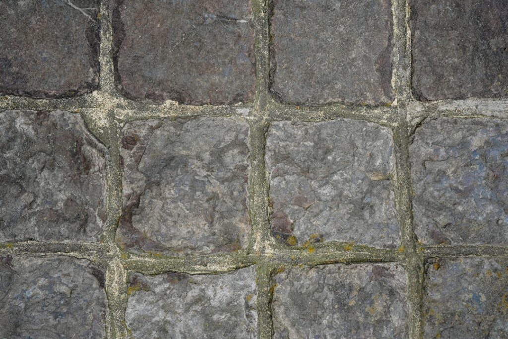 Close up of a gray stone cobblestone wall with mossy mortar lines between square blocks