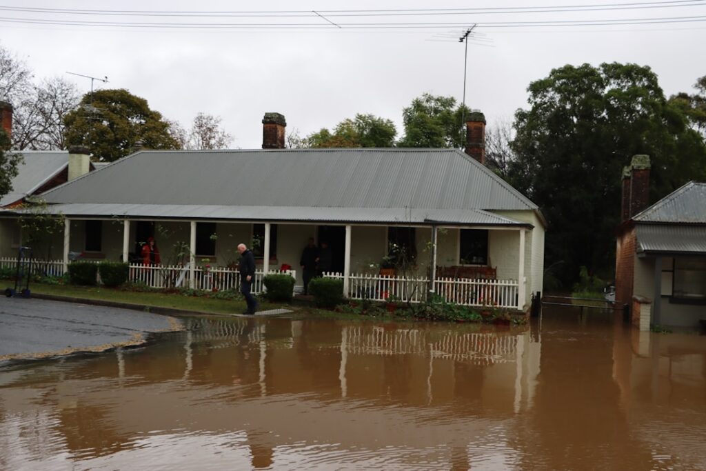 A single story house with a metal roof sits beside floodwater that reaches the yard two people stand on the front porch