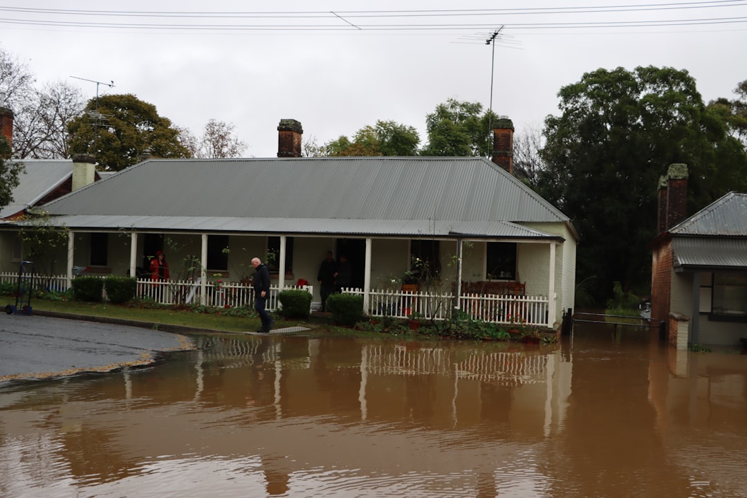 A single-story house with a metal roof sits beside floodwater that reaches the yard; two people stand on the front porch."