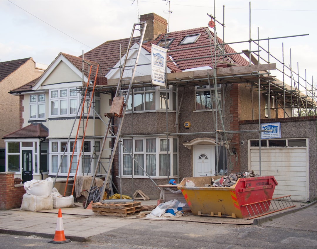Two-story house undergoing renovation with scaffolding along the façade, ladders, and a yellow rubble skip outside the front yard.”