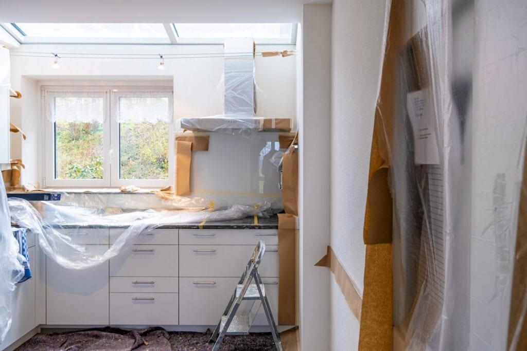 Kitchen under renovation with plastic coverings, taped surfaces, and a step ladder by white cabinets beneath a window.