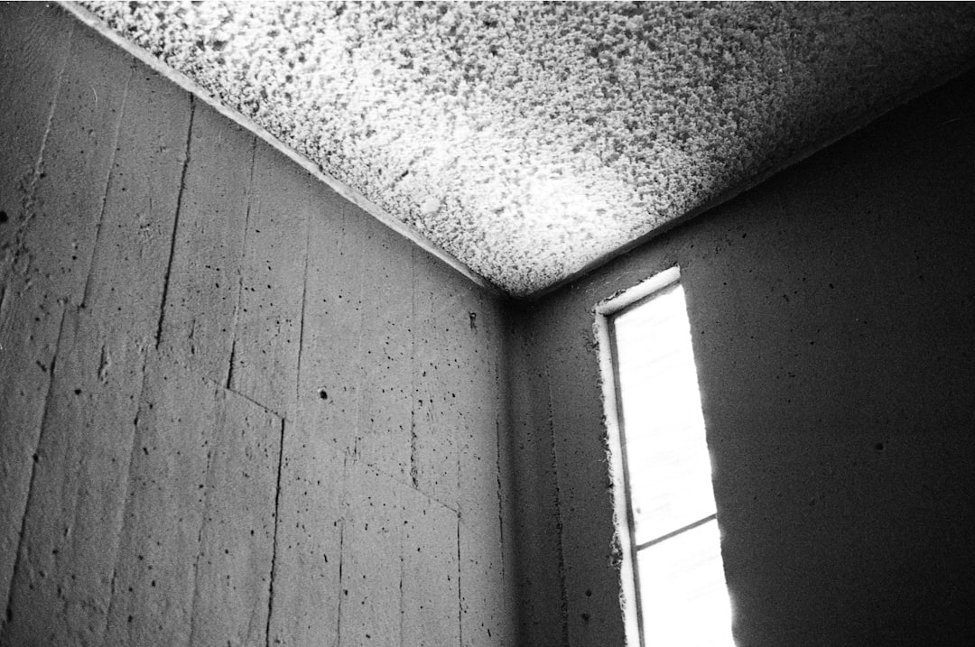 Black-and-white photo showing a bare corner of a room, with a textured ceiling and a tall narrow window letting light spill in.