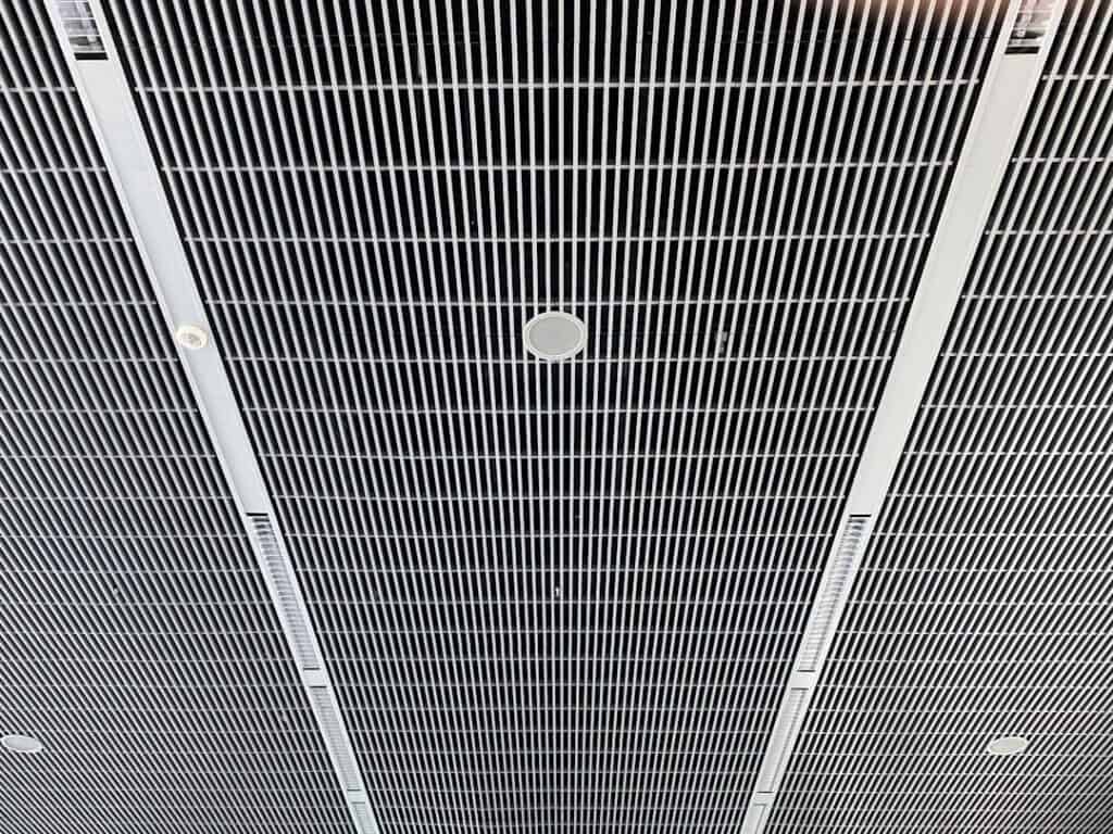 Close-up of a suspended metal grid ceiling with parallel slats and circular recessed speakers or vents scattered throughout the grid.