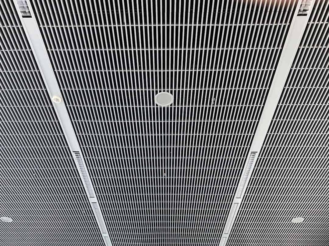 Close-up of a suspended metal grid ceiling with parallel slats and circular recessed speakers or vents scattered throughout the grid.
