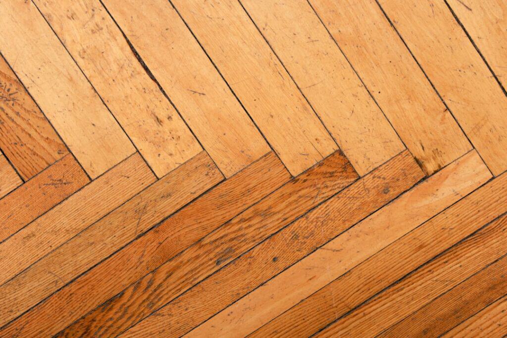 Diagonal wooden floor with warm brown planks arranged in a herringbone pattern, showing grain and wear marks.