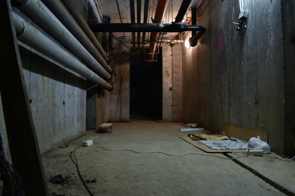 Underground utility corridor with exposed pipes along concrete walls and a single light fixture.