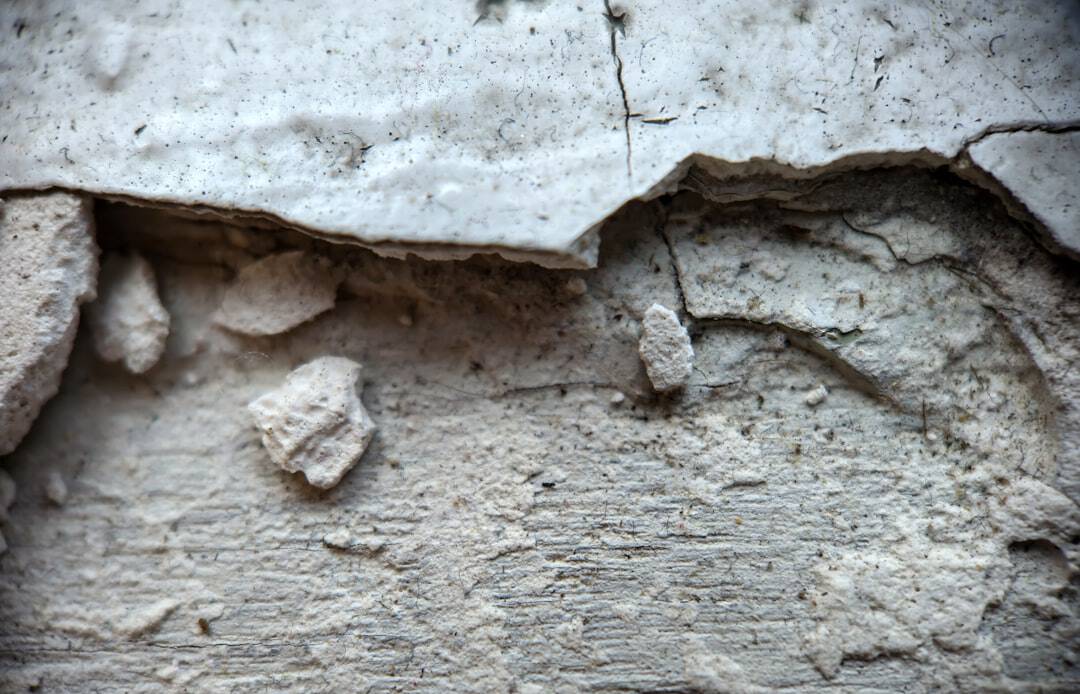 Close-up of a cracked, crumbling plaster wall with exposed concrete and dust scattered around the rubble نزدیک.