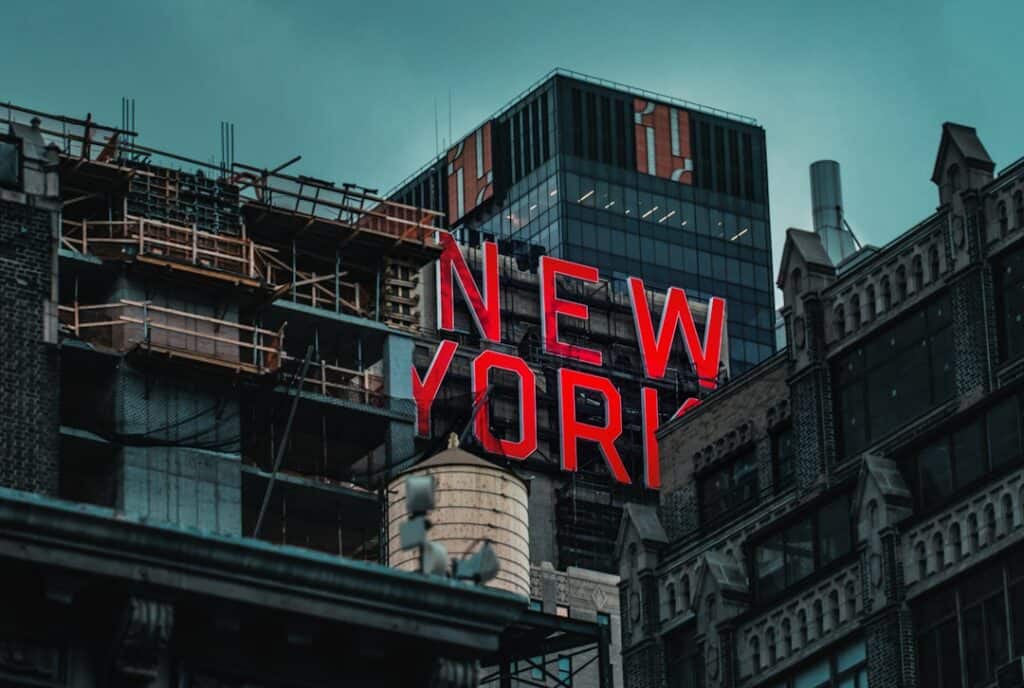 Red neon 'NEW YORK' sign atop a city building with construction scaffolding on the left and modern glass towers in the background.