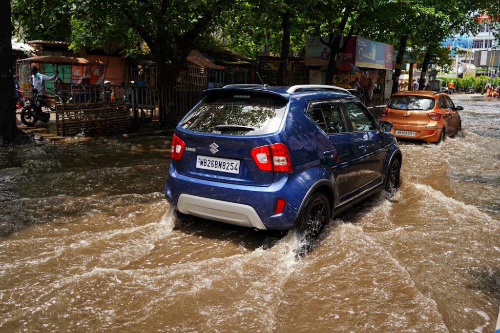Blue compact SUV driving through muddy floodwater on a busy street, with market stalls and pedestrians in the background.
