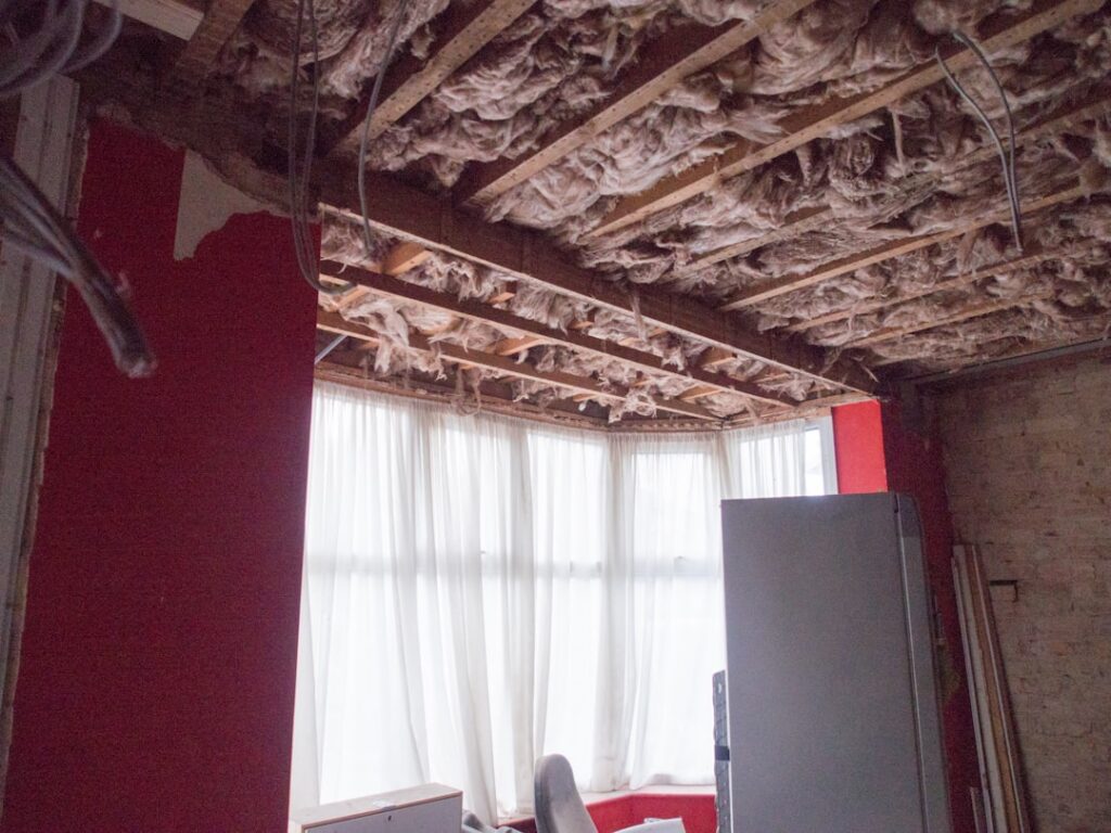 Room under renovation with exposed insulation in the ceiling, red walls, and a large window with sheer curtains; a tall gray refrigerator sits near the corner.